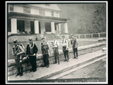 Six men with equipment (mine rescue corps) standing on the sidewalk in front of the clubhouse. This picture is also found in the DeHaven Collection, Roll 1602 04.