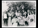 Fourth of July picnic at "Holmfield" at Nuttall Mountain by members of the Taylor family. Group of men, women, and children in front of a tree. United States flags included. <p> L-R standing: Jackson Taylor, Florence Field, Andrew Taylor, Fred Raven, Minnie Raven, John Taylor, Martha Taylor (baby), Jerry Taylor, Minnie Foster, Maria, and Alice Taylor. L-R front: May Rothwell, Florence Herrington, Tom Taylor, Mira Mercer, and Marcie Taylor (Hines). <p> Identifications by Marcie T. Hines in 1954: All of the Taylors including Martha (the baby). Mira Mercer her mother at Tom's left. May Rothrocle, John's second wife, at Tom's right. Florence T. Herrington, daughter of William Taylor of Newburg, NY, back of Tom. Minnie Foster, daughter of William Foster of Newburg, NY, in front of Alice and Jerry. A friend, Florence Field, standing by Marcie Hines's father. The top of Maria's head visible in back of Alice. Marcie Hines draped in a flag. Minnie and Fred are in the middle, Andrew behind May.