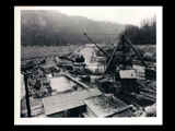 East end of dam during the hydroelectric power construction on the New River. View shows upstream retaining wall and upstream end of downstream retaining wall. Excavation for east abutment between these walls shown flooded out. Block No. 3 of dam shown in foreground with temporary railroad crossing for concrete cars. New-Kanawha Power Company, Hawks Nest - Gauley Junction Development No. 214.