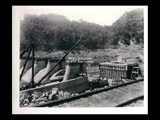 Dam during the hydroelectric power construction on the New River. View of east end of dam showing downstream retaining wall, east abutment and upstream retaining wall. When the abutment section is completed, retaining walls shown will be backfilled. Men working. Buildings alongshore in the distance. New-Kanawha Power Company, Hawks Nest - Gauley Junction Development No. 253.