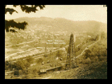 View of Mannington from hillside. Oil derrick in foreground. Traction line, railroad tracks and cars.