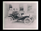 Two Wheeling policemen in automobile on brick street in front of building with two men looking on from window.