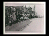 Group of people waiting at a streetcar island stop on brick (Main?) street. Signs include M H & M Shoes Hosiery, The Children's Shop, George Hill Dentist.