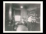 Interior view of Ohio Valley Hospital showing the pharmacy with a woman sitting at a desk and a man standing at a counter with drug jars on shelves.