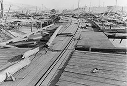 This view looks south from the munitions pier, showing the wreckage of Building A-7 (Joiner Shop) at the right. There is a piece of twisted steel plating just to the left of the long pole in the left center. Photo was taken by the Mare Island Navy Yard (NH 96821).