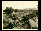 Footers on Building 2 (East Wing) during construction of the Cass Gilbert WV State Capitol building. A railroad car is at right, houses in the background.