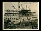 Second level stonework on Building 2 (East Wing) during construction of the Cass Gilbert WV State Capitol building. Girder structure visible above. Railroad cars are in the foreground.