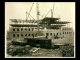 Second level stonework on Building 2 (East Wing) during construction of the Cass Gilbert WV State Capitol building. Girder structure visible above. Railroad cars are in the foreground.