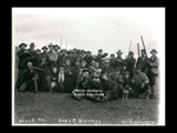 Rabbit hunters on McCanns Run. A group of men, women, and children are posed outside, many holding guns in their hands.  Two dogs are with the group.