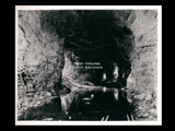 View into tunnel during the hydroelectric power construction on the New River. View shows tunnel roughly excavated to 32 foot diameter with sandstone roof and approximately lower 2/3 of tunnel in shale. Final trimming and invert excavation have not been started. The adit to surge basin site is shown at left. Water in tunnel floor. Camera 9 feet right of centerline and at Station 89 + 70. New-Kanawha Power Company, Hawks Nest - Gauley Junction Development No. 326.