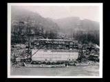 Power house during the hydroelectric power construction on the New River. View taken from across the river showing substructure and superstructure steelwork completed. Buildings on the hillside behind. New-Kanawha Power Company, Hawks Nest - Gauley Junction Development No. 347.