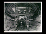 View into tunnel during the hydroelectric power construction on the New River. View looking downstream showing the combination scaling car and concrete gun carriage. The concrete hopper is mounted immediately over the gun on the right hand side of the carriage. The traveling steel formwork is shown partially assembled beyond the scaling car and gun carriage. Camera at Station 21 + 80 and scaling car and gun carriage at Station 22 + 20. New- Kanawha Power Company, Hawks Nest - Gauley Junction Development No. 362.