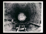 View down tunnel during the hydroelectric power construction on the New River. View shows alternate portions of concrete inverts completed and excavation finished for remaining inverts. Templates are shown on each side of the unfinished invert for the bolts which hold the overlapping portion of the steel form to the finished invert concrete when placing the tunnel lining. Camera at Station 84 + 50 looking downstream. New-Kanawha Power Company Hawks Nest - Gauley Junction Development No. 366.