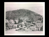 Looking east at buildings in Hundred with tree covered hill behind.