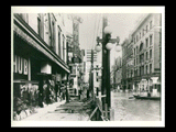 View down street during 1913 Wheeling flood near intersection with 14th Street. Merchants built a minature dike to protect their businesses. Signs include The Hub, House & Herrmann, Kurutz Brothers, American Restaurant, Baumer, pianos, Victrolas, carpets, furniture, "Do Not Spit on Sidewalk."