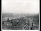 Aerial view of Wheeling. Signs for Chas Elig Carriage Co., John S. Welty and Sons Wholesale and Retail Grocers, and W. H. Colvig Millinery. Suspension bridge at far right.